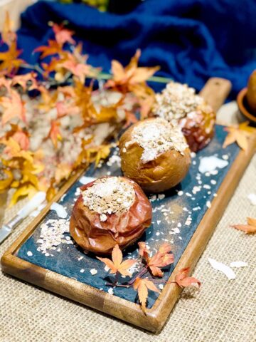 Three baked apples with oatmeal on a blackboard with a wooden frame dusted with cinnamon and autumnal leaves for decoration.