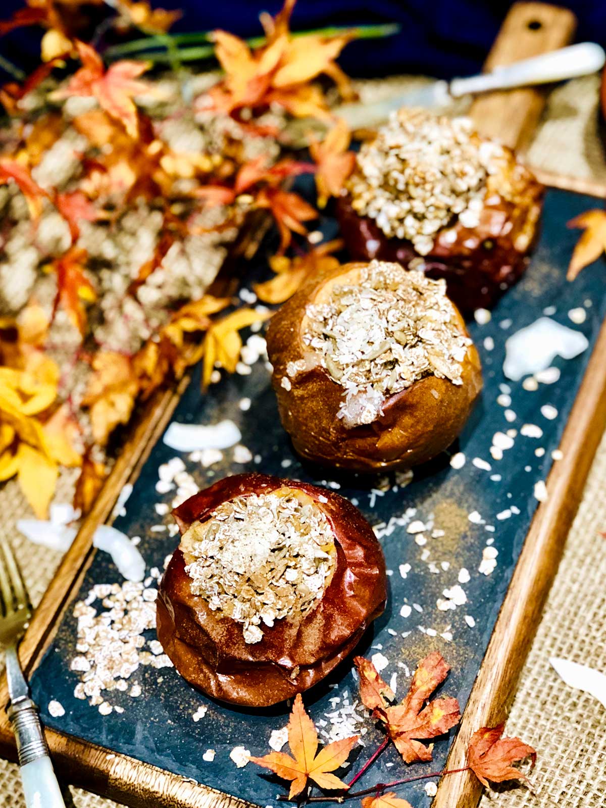 Close up or birdseye of three baked apples stuffed with oatmeal and coconut flakes sprinkled over on a black and wooden frame board.