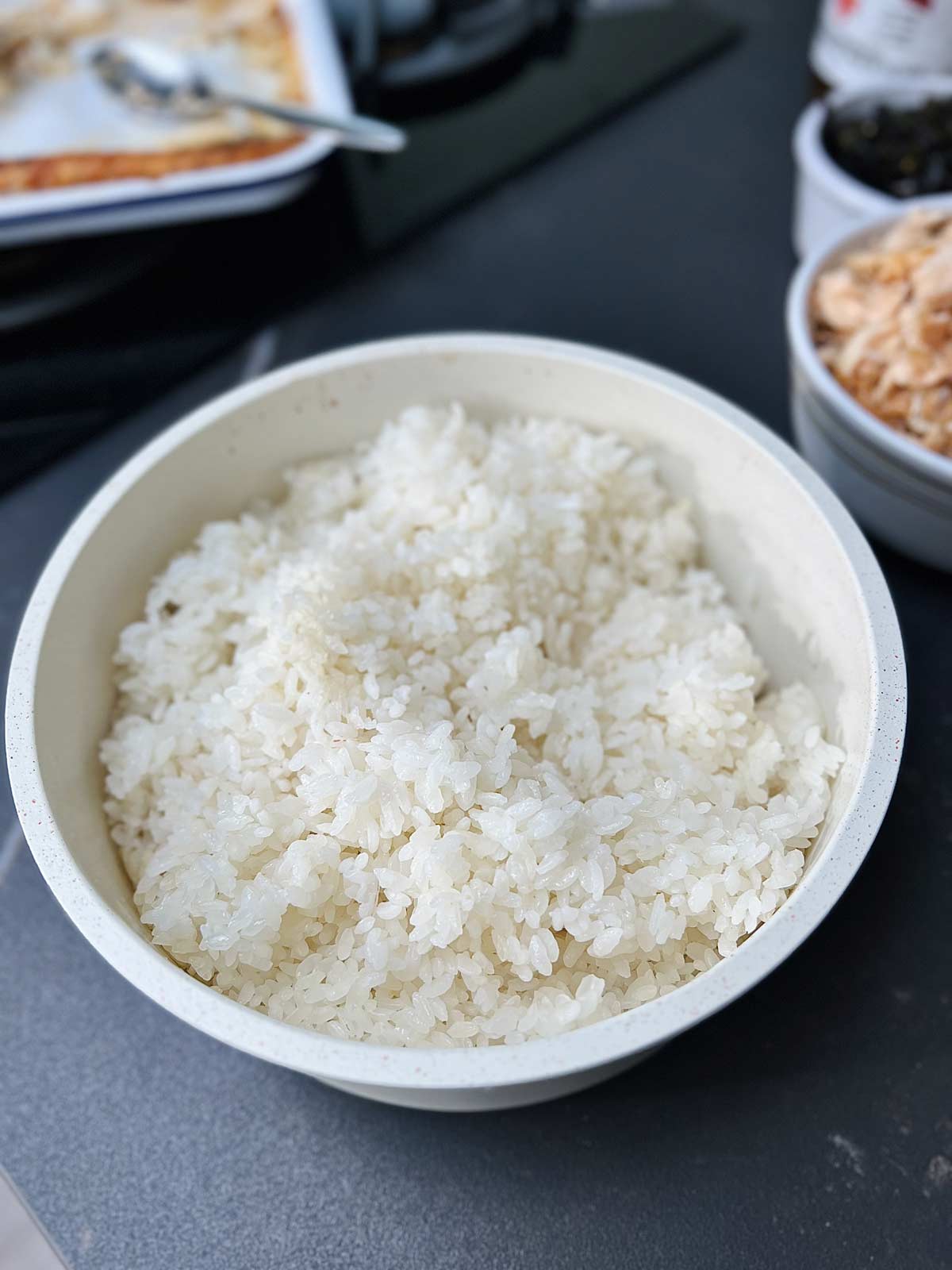 Boiled sushi rice in a white dish on a dark surface.