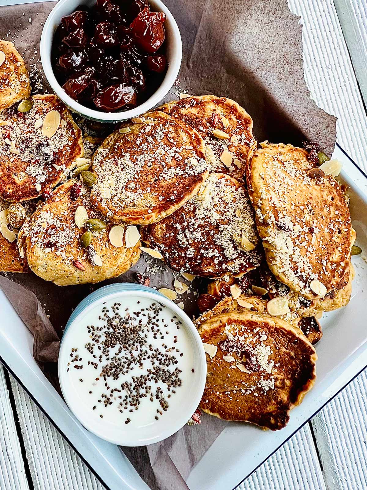 Birds eye view of butternut squash pancakes sprinkled with almonds, almond flour, pumpkin seeds, goji berries and two small serving dishes with cherry jam and yogurt and chia seeds on a white metallic tray