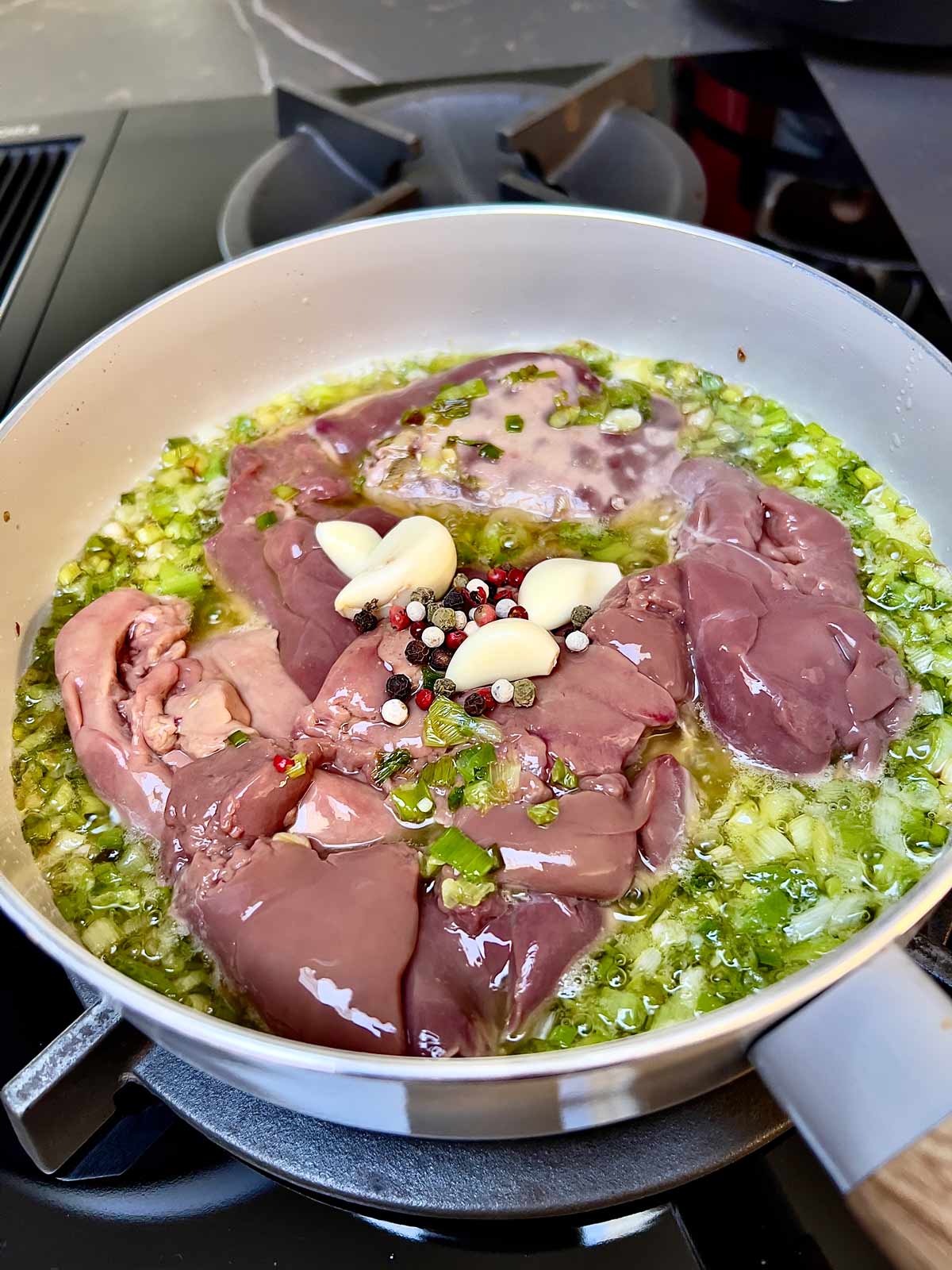 Chicken livers added to a pan that contains cooked scallions with four gloves of garlic and mixed peppercorns.