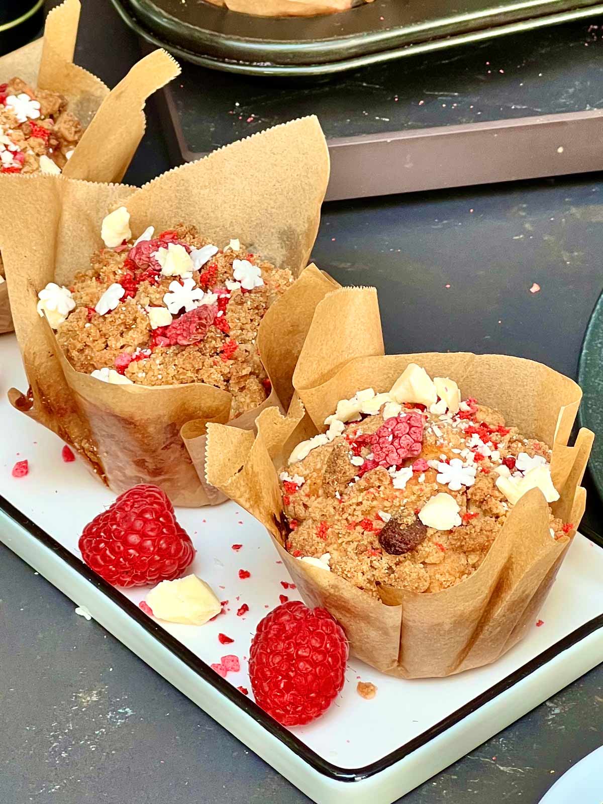 Close up of two white chocolate raspberry muffins sprinkled with crumbs of frozen dried raspberries and white chocolate on a white rectangular ceramic plate.