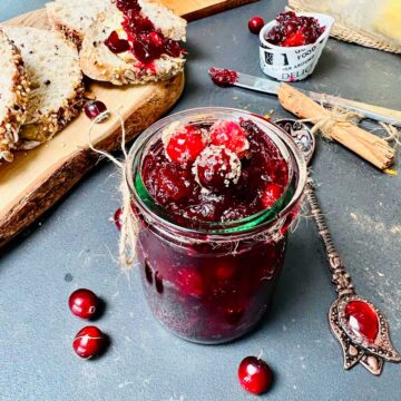 Cranberry jam in a little glass on a dark worktop, a wooden board with bread slices above and an artisan teaspoon right next to it