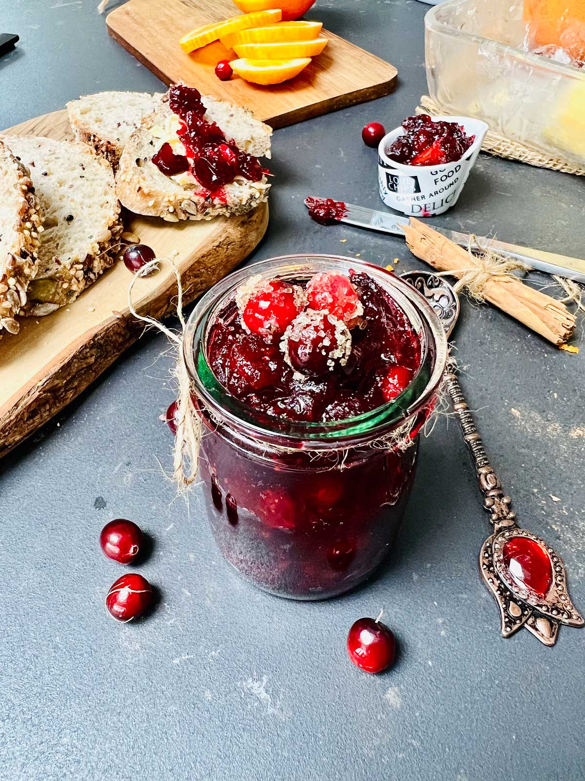 Cranberry jam in a little glass on a dark worktop, a wooden board with bread slices above and an artisan teaspoon right next to it