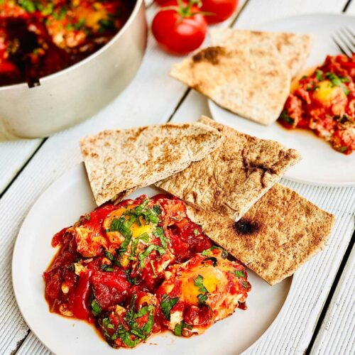 Shakshuka on two white plates with pieces of flatbread on the edge of the plates and a bunch of fresh tomatoes at the top middle of the picture and a glimpse of the pan with shakshuka in on the top left corner of the picture