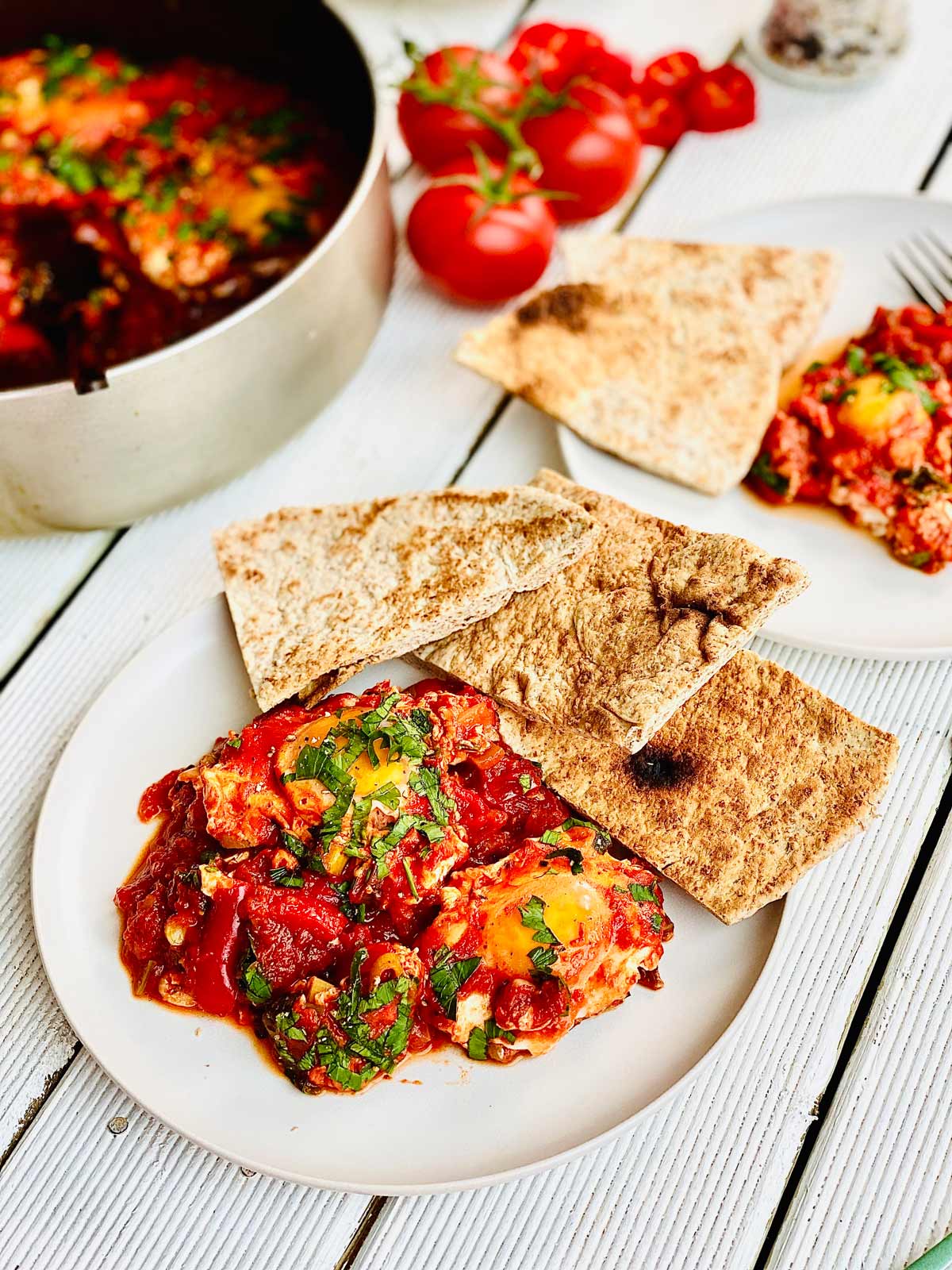 Shakshuka on two white plates with pieces of flatbread on the edge of the plates and a bunch of fresh tomatoes at the top middle of the picture and a glimpse of the pan with shakshuka in on the top left corner of the picture