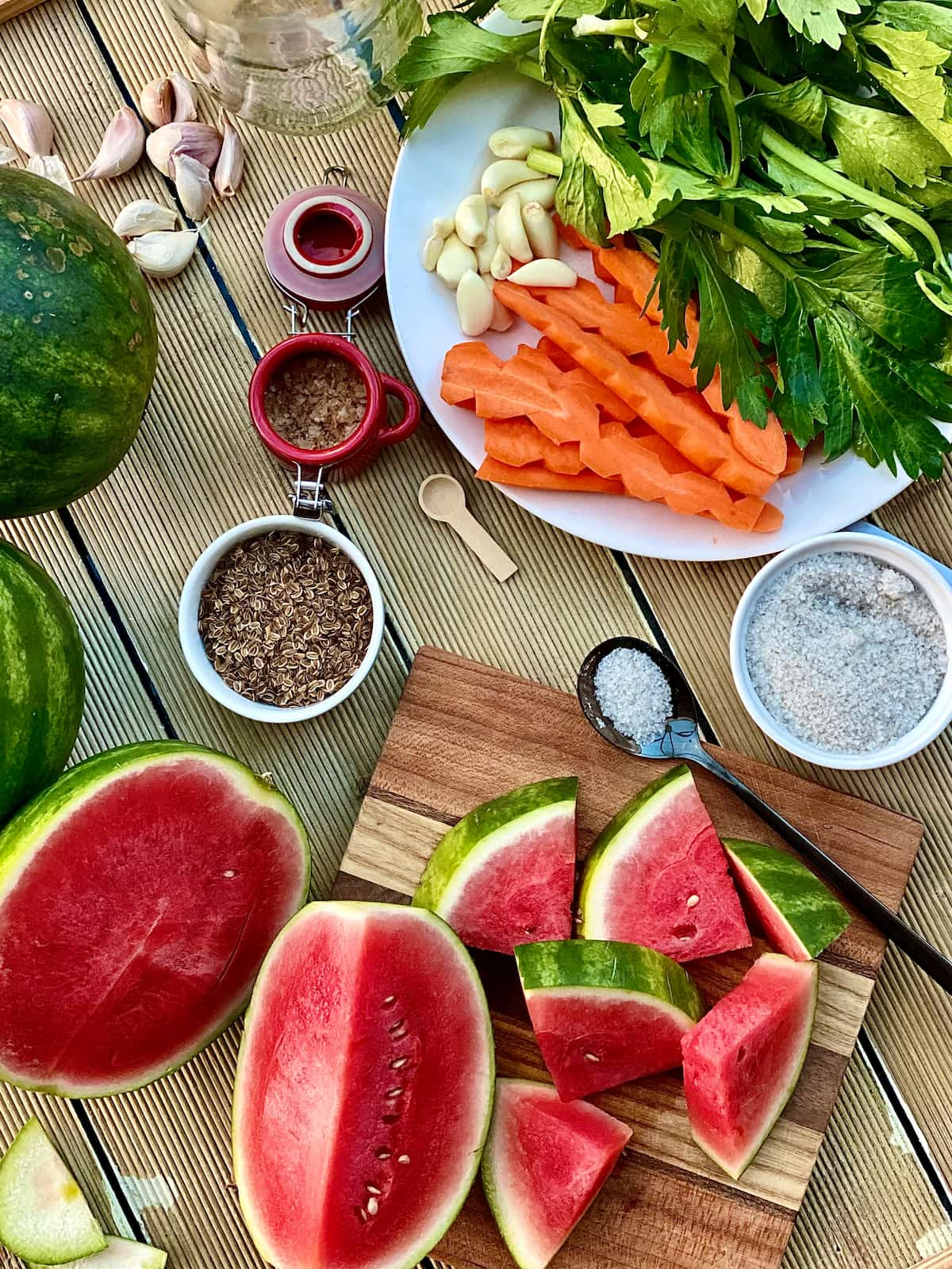 Fresh watermelon sliced and quartered for pickling, salt in a tiny dish, dill seeds and carrot, garlic cloves, celery leaves, on a white plate