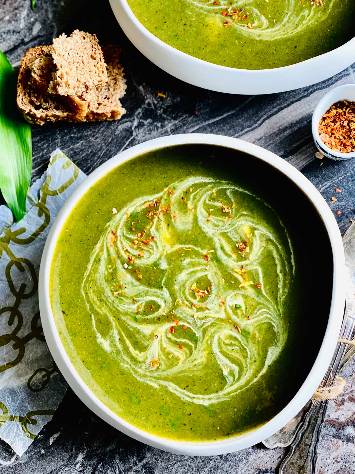 Freshly foraged wild garlic soup in a white presentable soup bowl birds-eye view with swirled up cream and a pinch of chilli flakes on a dark black marble worktop