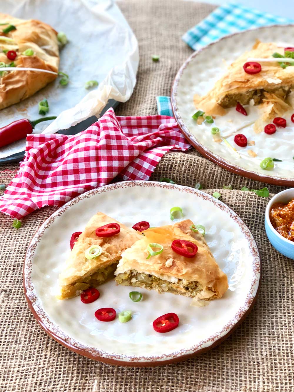 Giant potato and cauliflower filo samosa slices on a plate topped with chillies and chopped scallions.