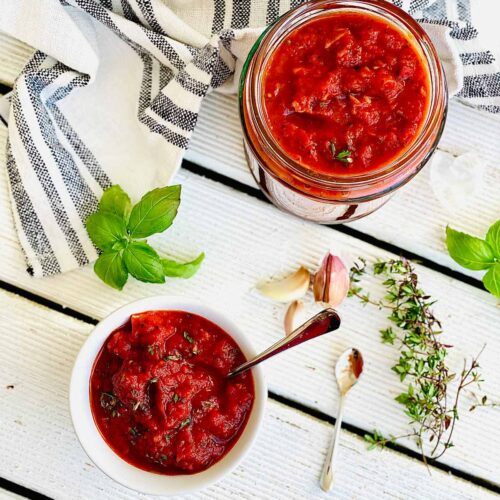 Homemade pizza sauce in a jar and in serving dish with herbs and garlic scattered around