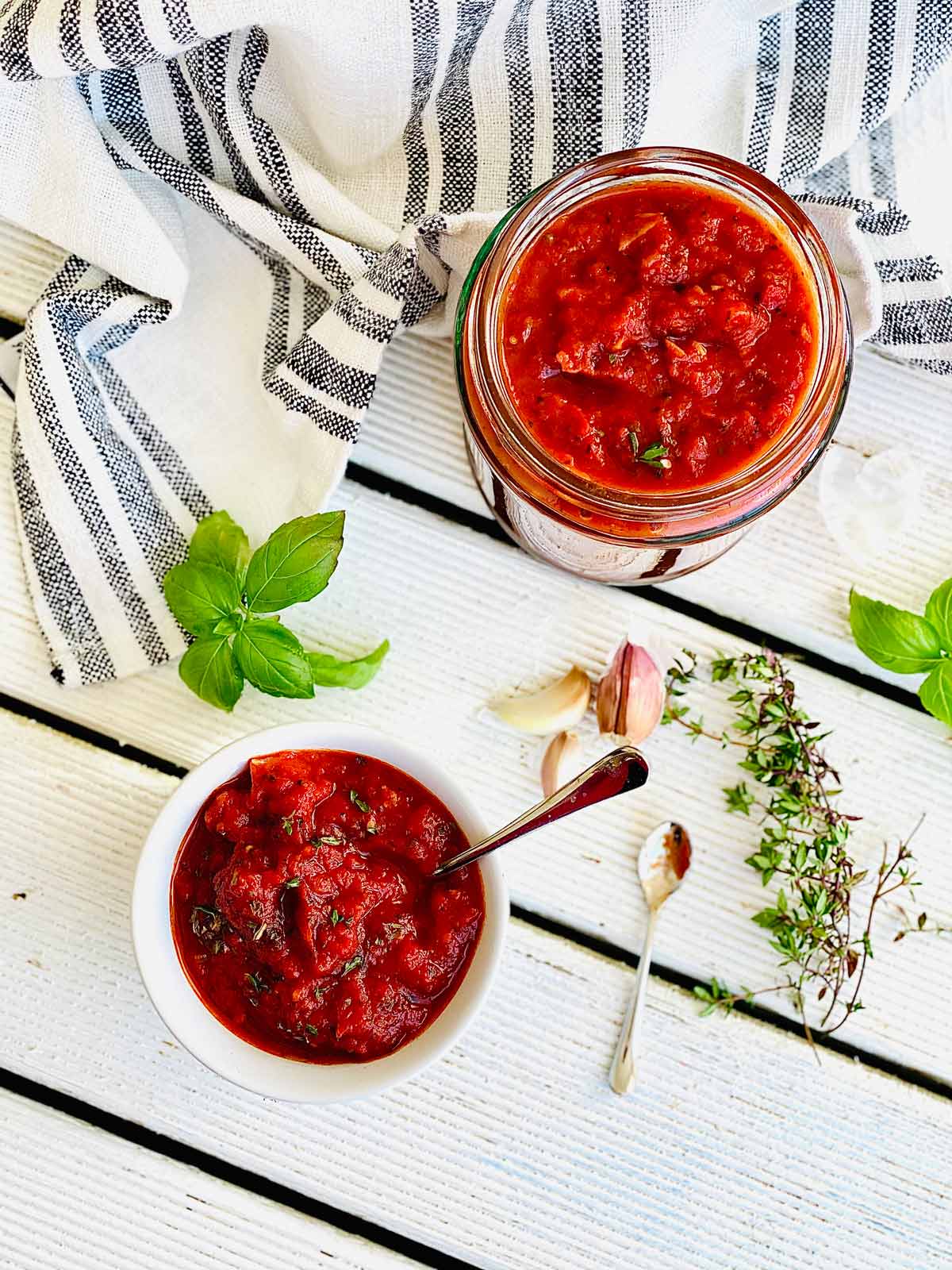 Homemade pizza sauce in a jar and in serving dish with herbs and garlic scattered around