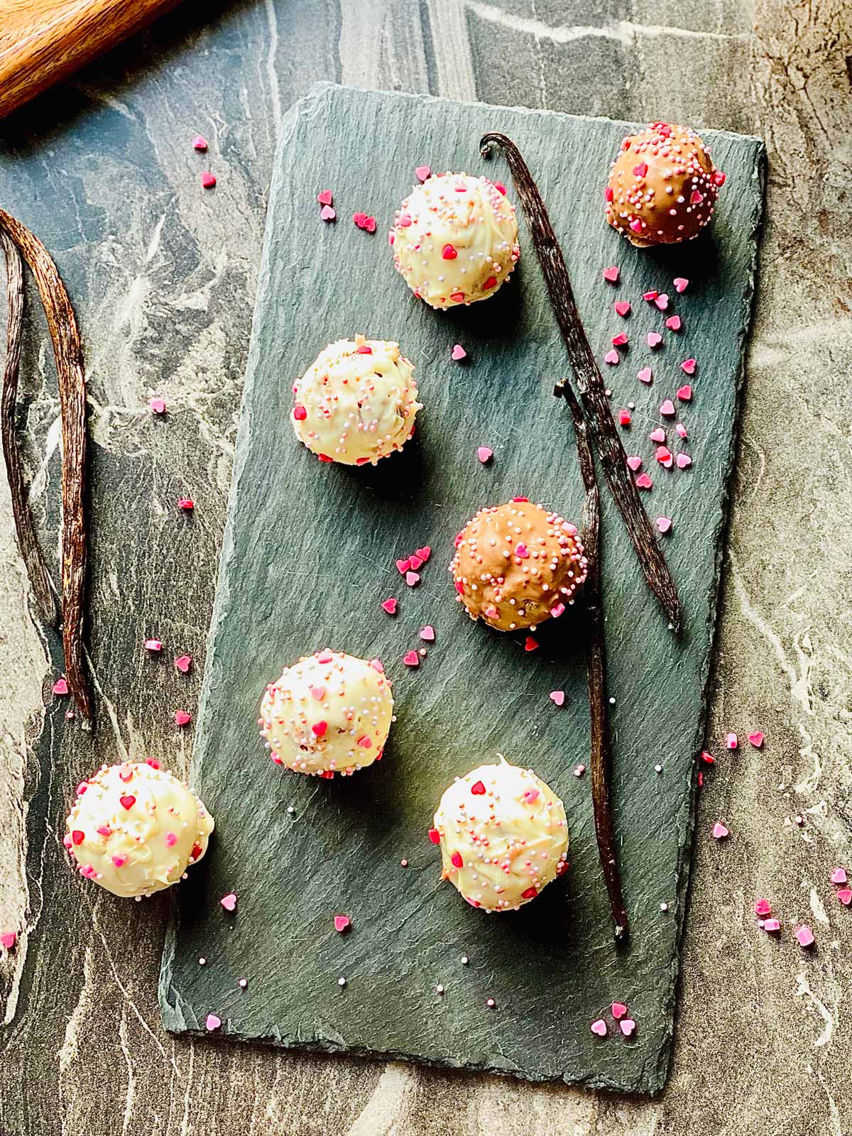 Cake pops on a black slate placemat with four vanilla pods and scattered little pink sugar hearts