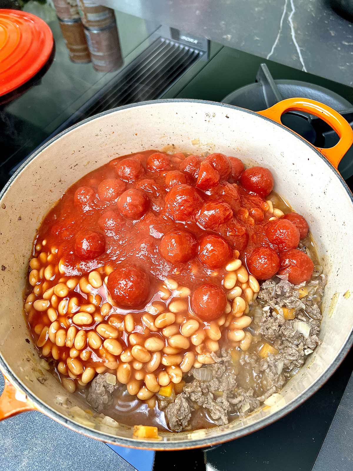 Minced beef half cooked with added baked beans and canned cherry tomatoes cooking in a deep pot.