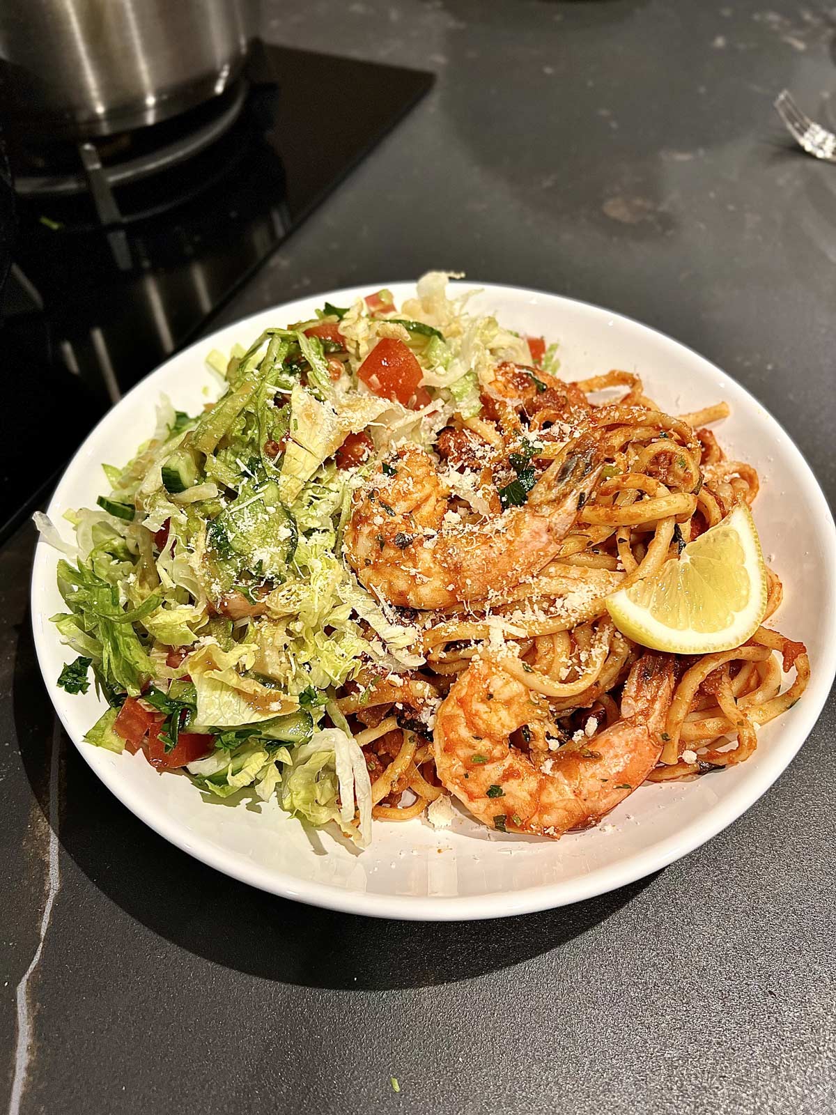 Prawn and chorizo, linguine pasta in a round white plate filled half with pasta and half with salad topped with a slice of lemon and a sprinkle of Parmesan.