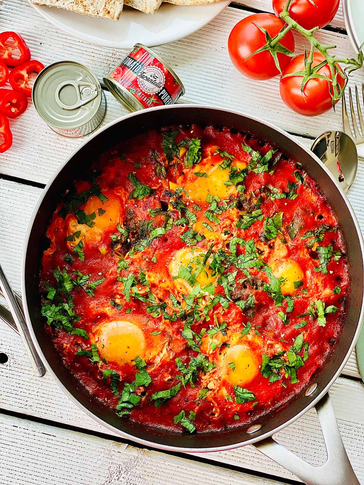 Shakshuka in a pan birds view with scattered fresh chopped parsley a couple of tiny tomato paste cans top left and some fresh tomatoes at the top right of the picture cutlery on the right