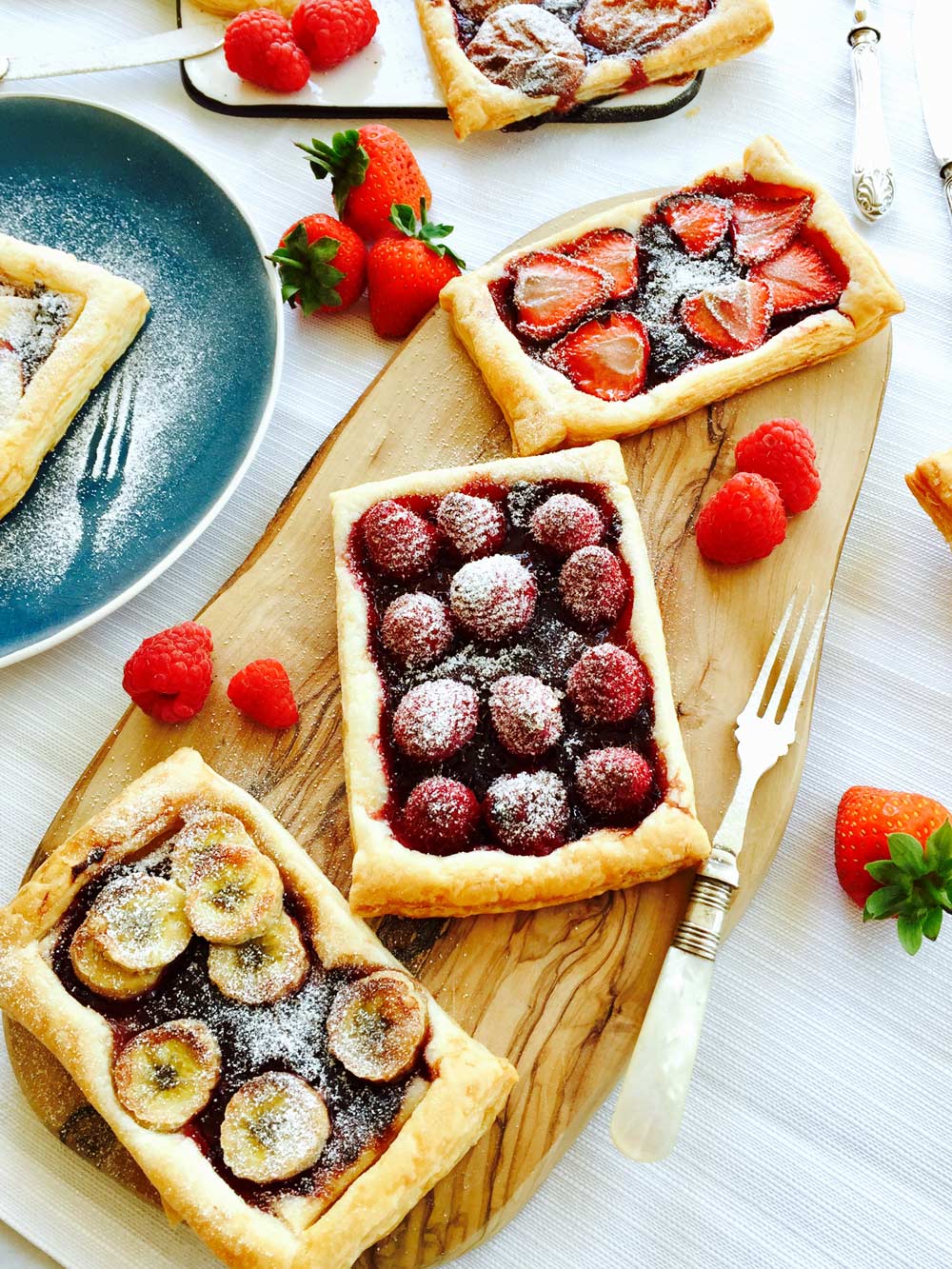 Three Tutti fruity jam and fruit rectangular tartlets on a wooden chopping board with fresh fruit scattered around.