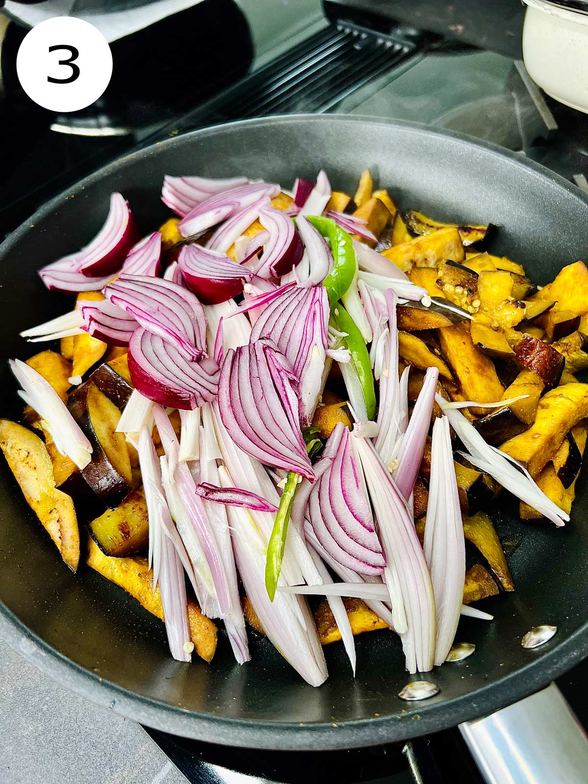 Aubergine chunks in a frying pan with wedges of onions and green chili added.