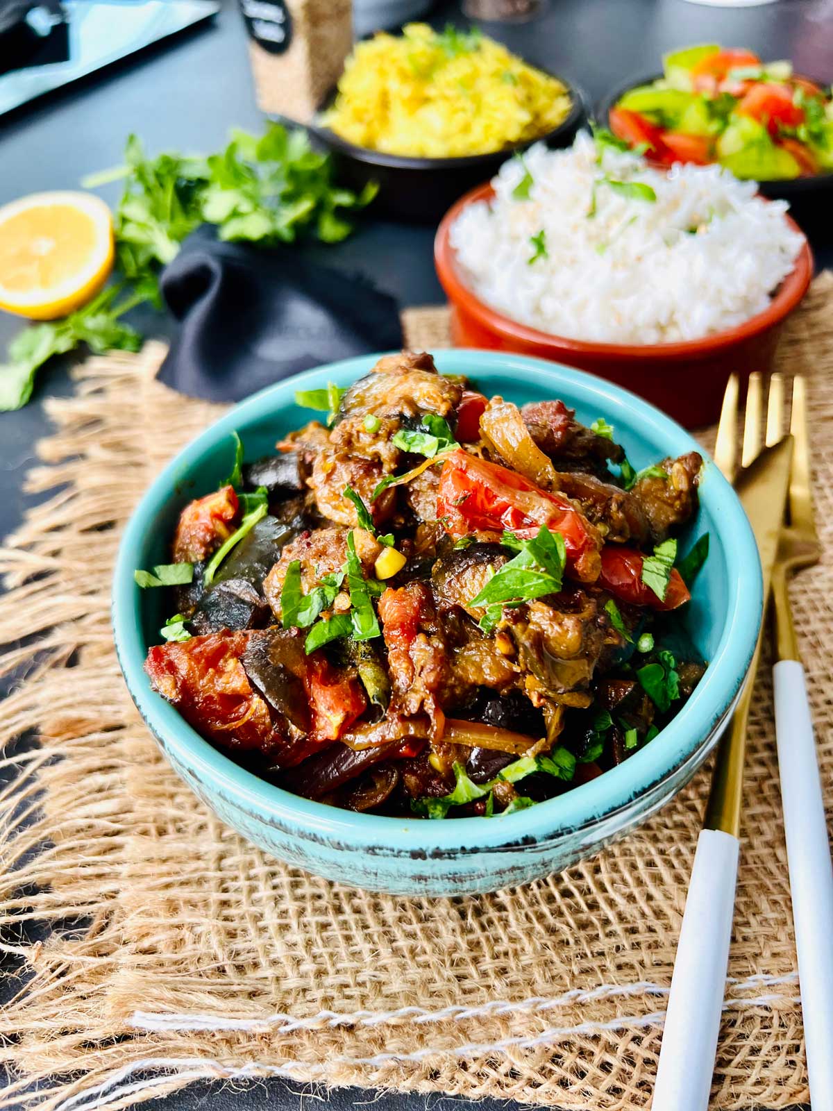 Aubergine curry in a ceramic pot served with rice with golden fork and golden knife to the side.