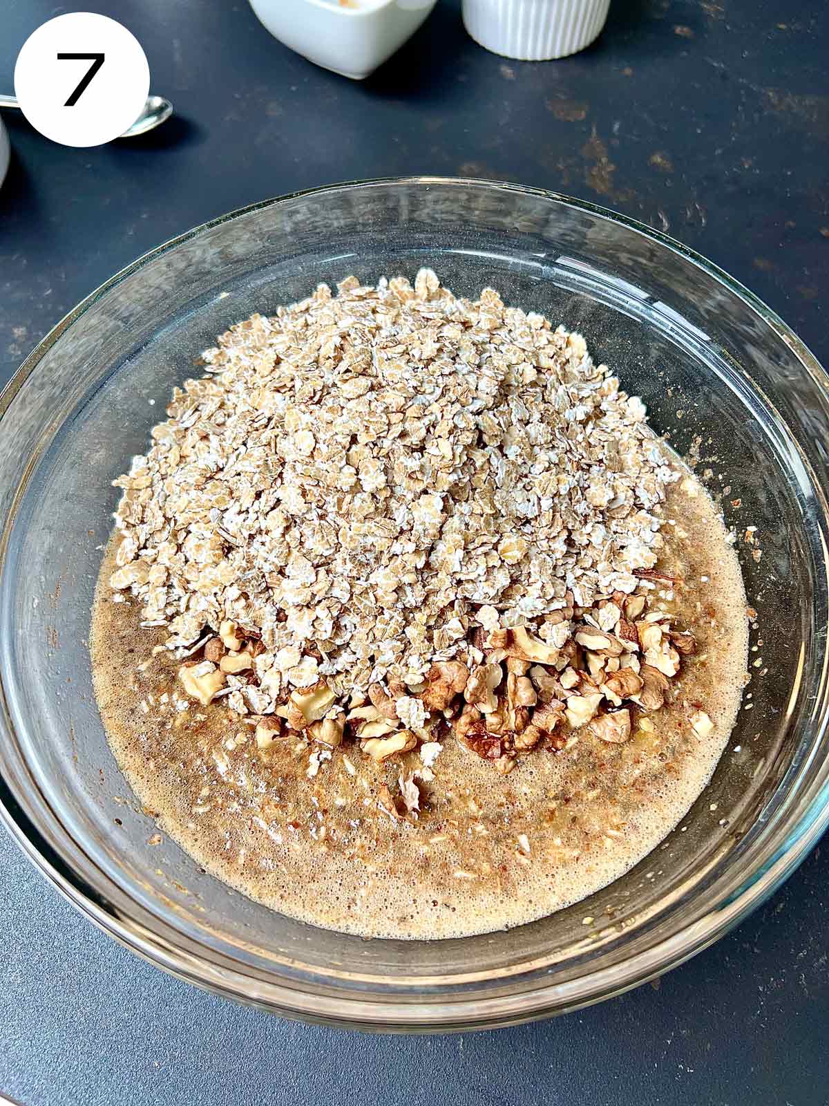 Banana bread mixture in a glass bowl with chopped walnuts and barley flakes added.