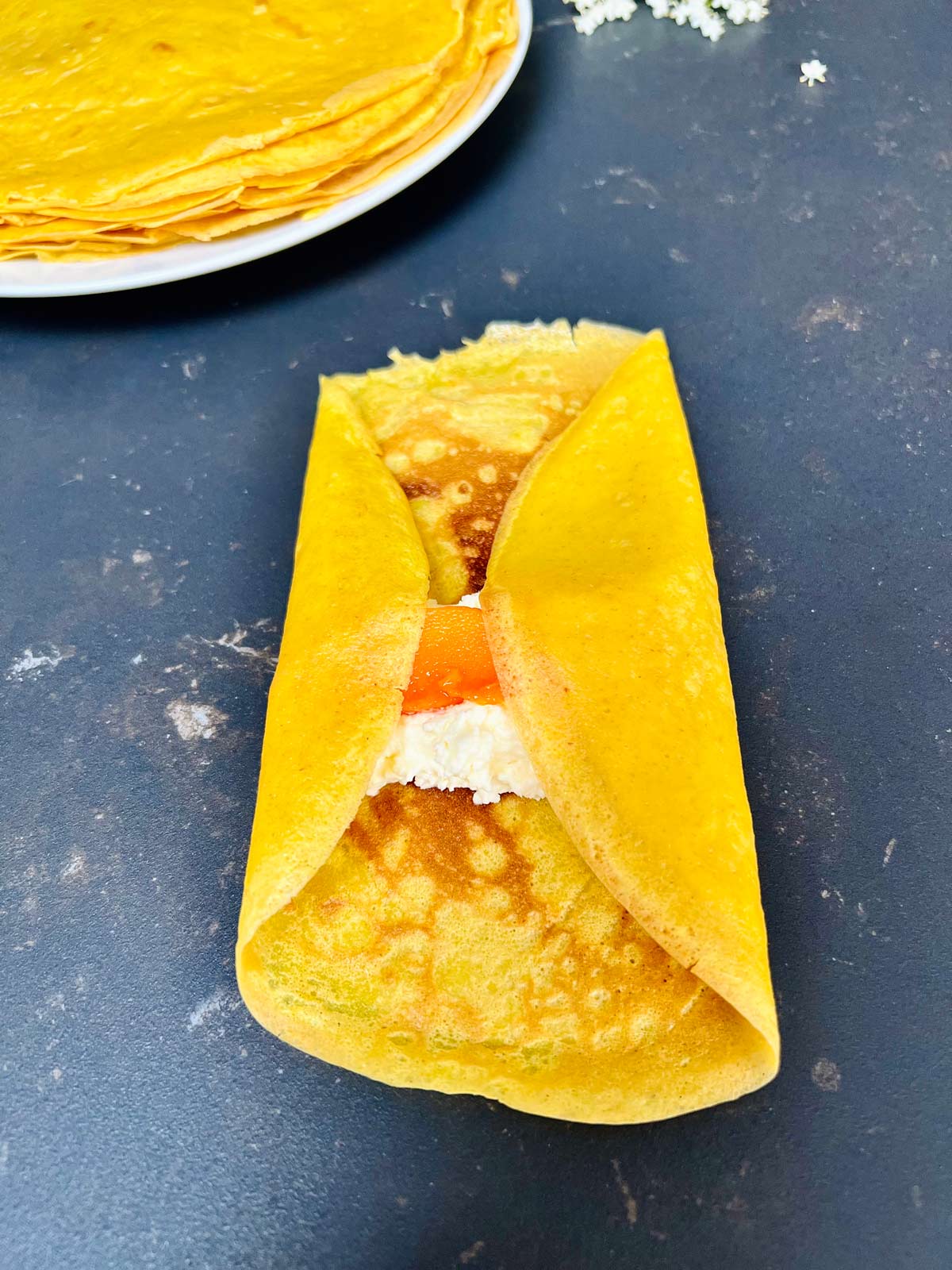 A bright yellow pancake, folded on both sides, with white filling and mango in the centre.