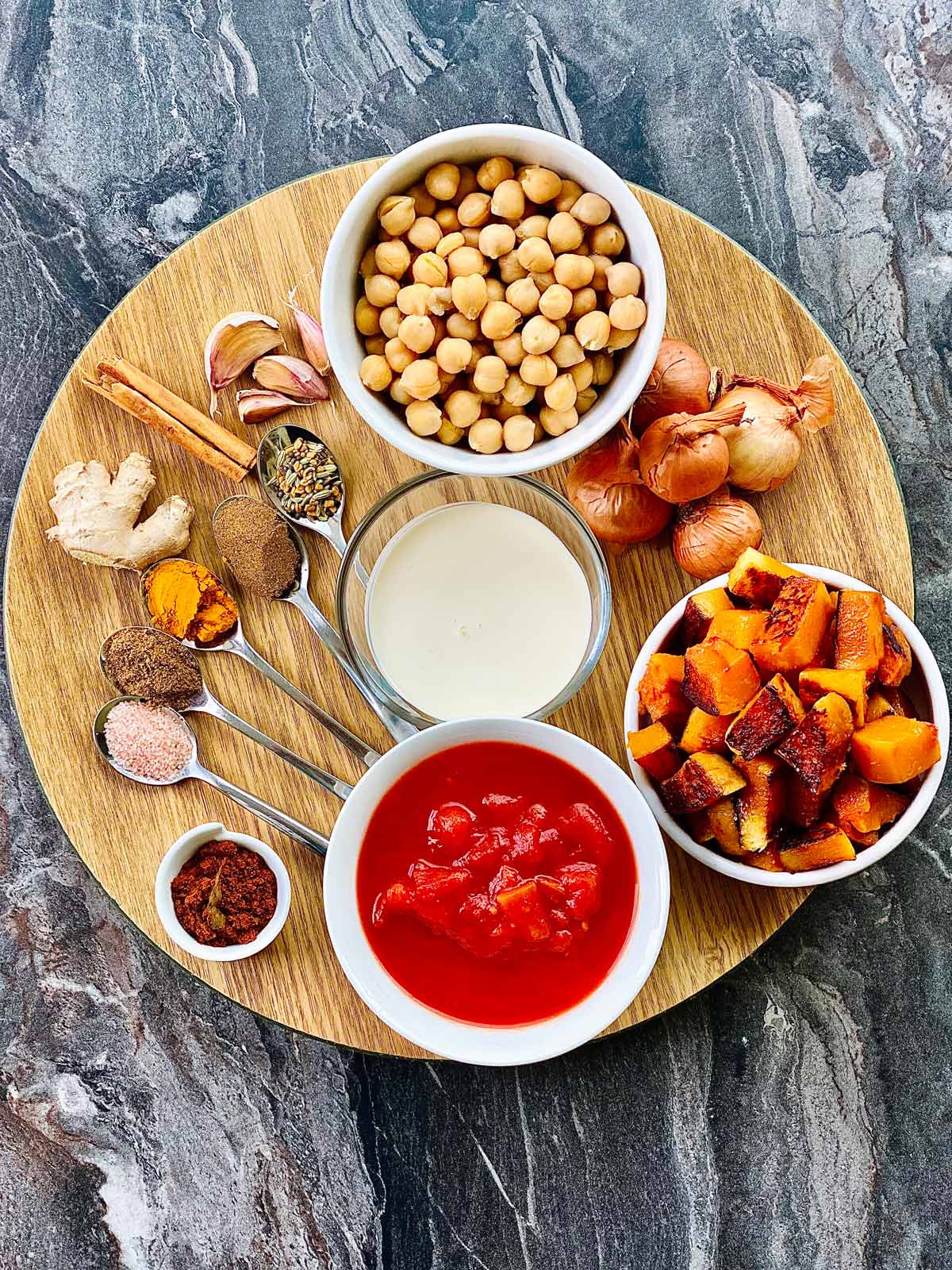 ingredients needed for butternut squash and chickpea curry on a round wooden plate - chickpeas, garlic cloves, 5 small onions / shallots spices on 5 teaspoons, ginger, cinnamon, butternut squash, curry powder, crushed tomatoes