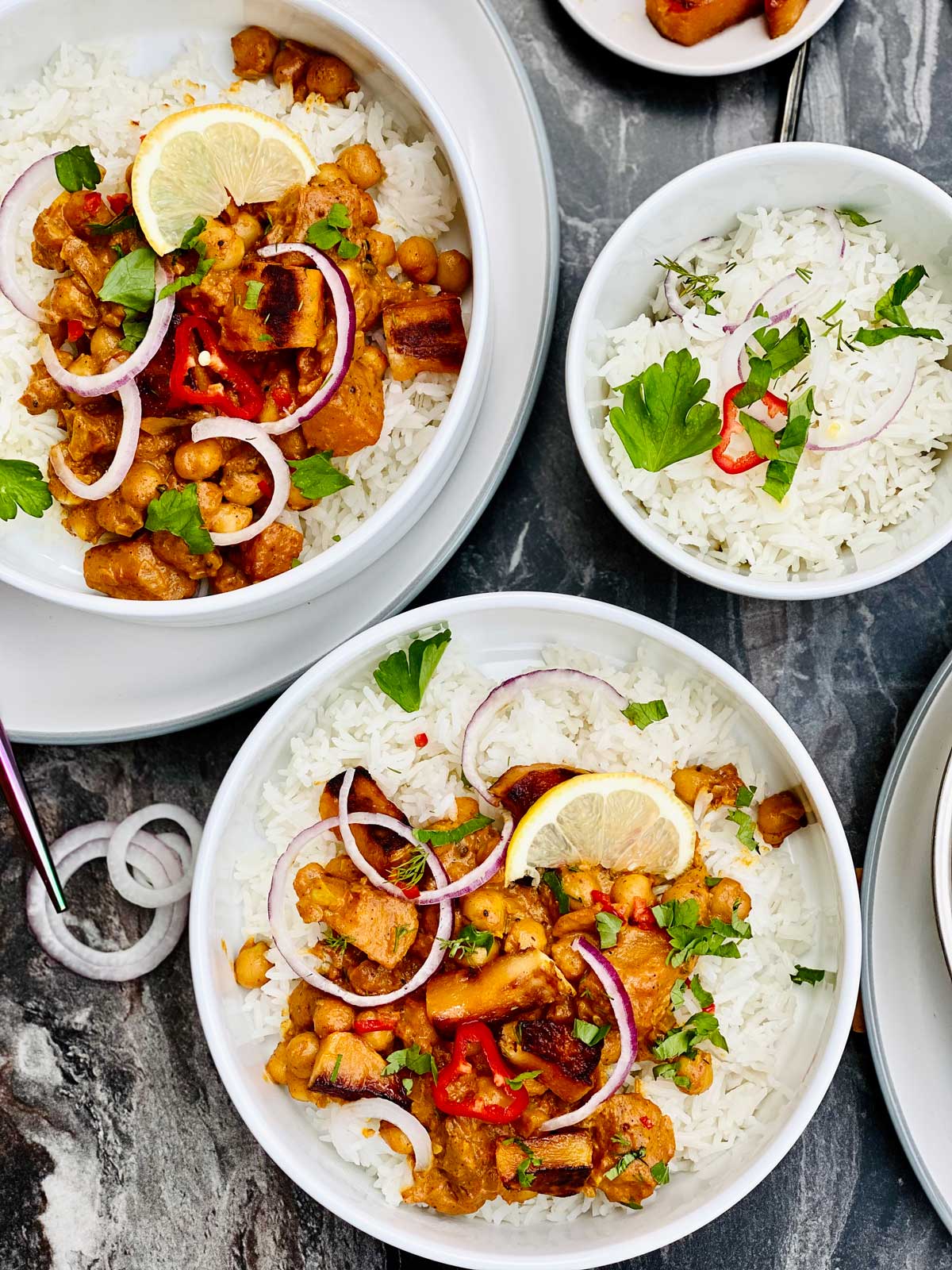 Birdseye view of butternut squash and chickpea curry on rice, served in white plates with a slice of lemon on top, fresh coriander scattered, rings of red onion and red chilli slices