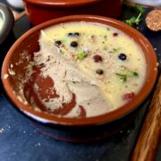 Chicken liver pate in a terracotta color dish topped with cooled butter, peppercorns and fresh herbs.