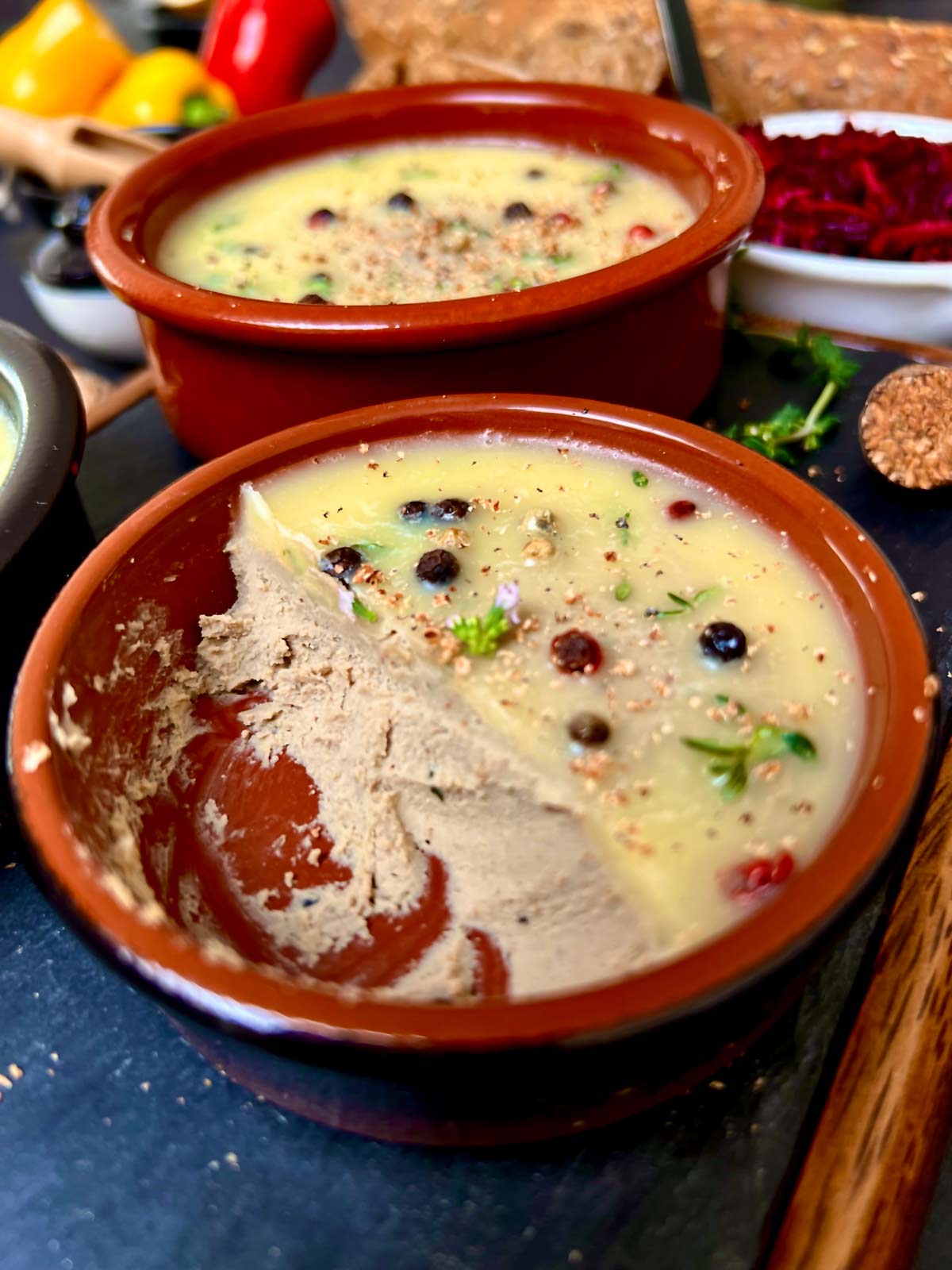 Close app of chicken liver pate in a terracotta color dish topped with cooled butter, peppercorns and fresh herbs.