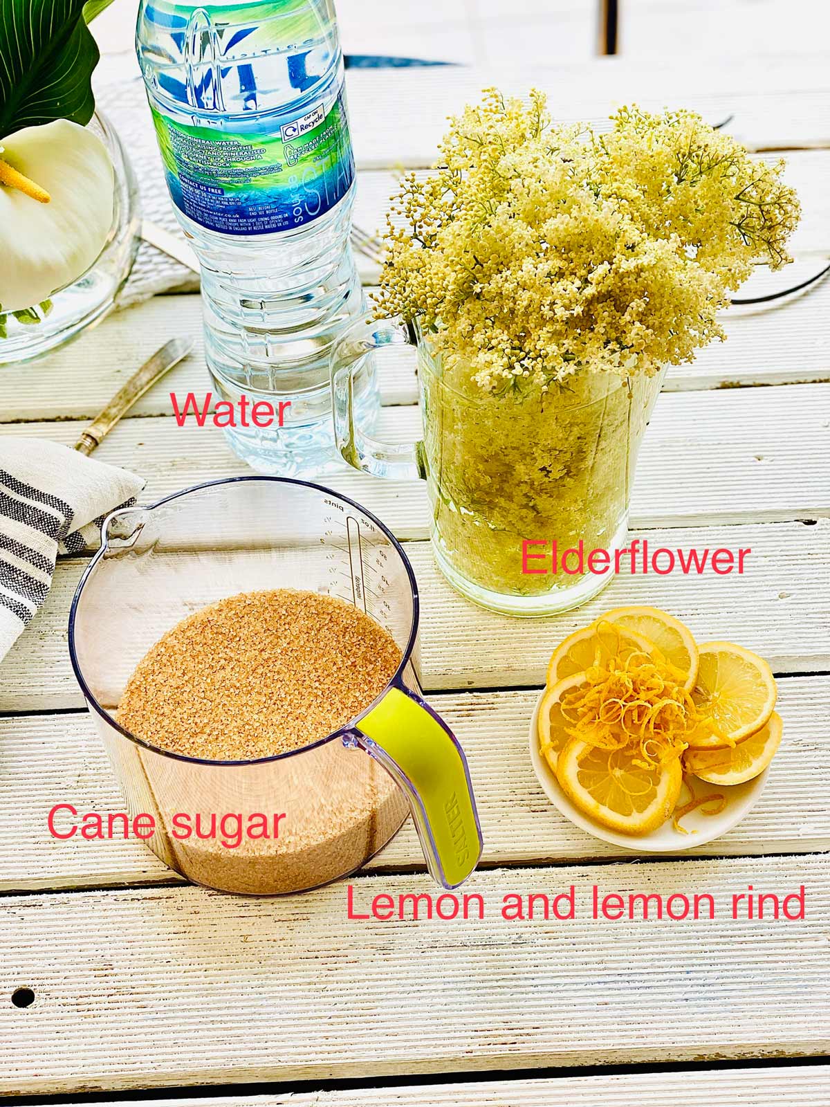 Ingredients needed for the elderflower cordial - cane sugar in a jug, lemon slices and lemon rind on the little white plate, elderflower in a tall jug and a bottle of spring water