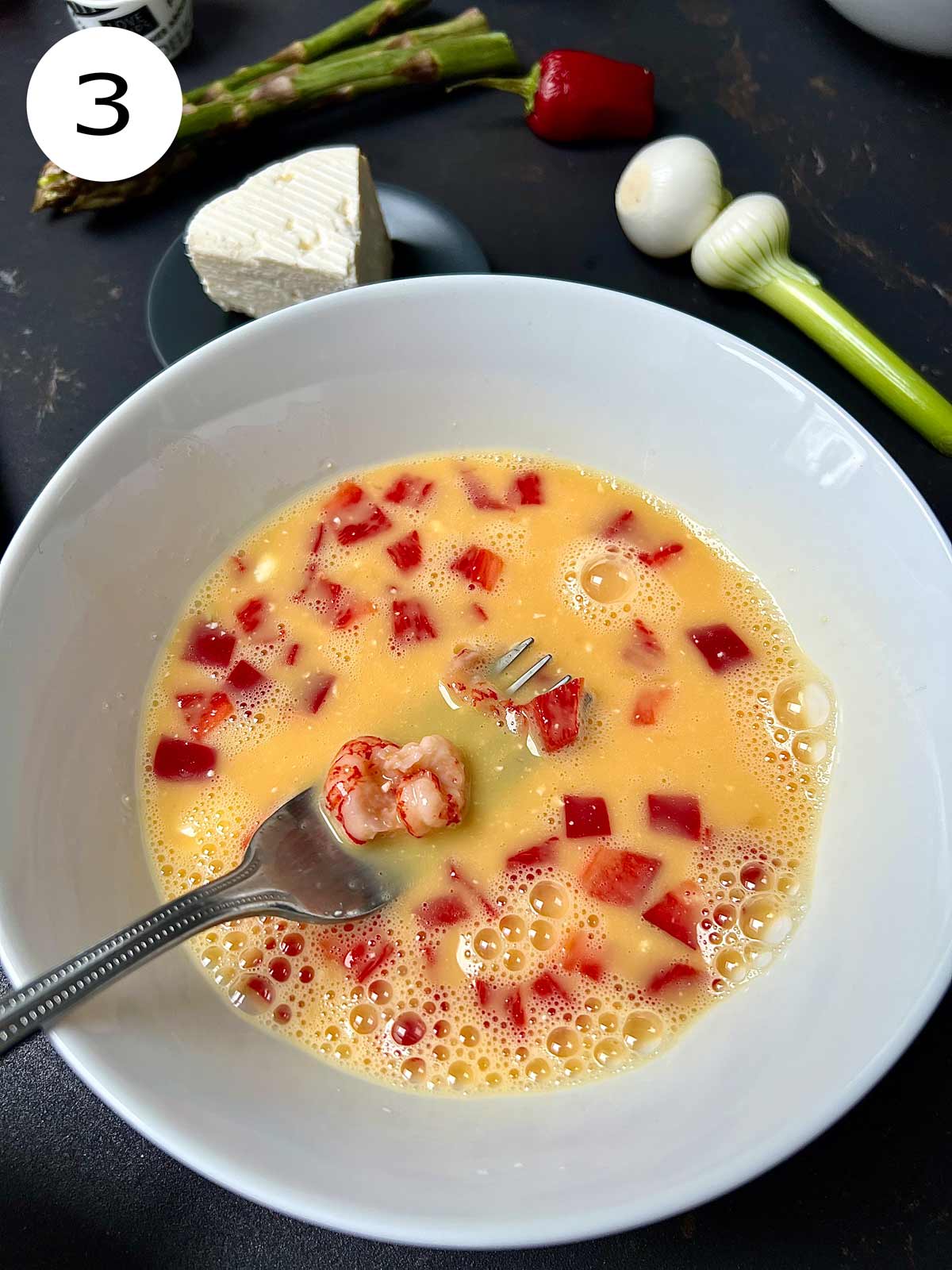 Eggs and finely chopped red pepper being beaten with a metalic fork in a white bowl.
