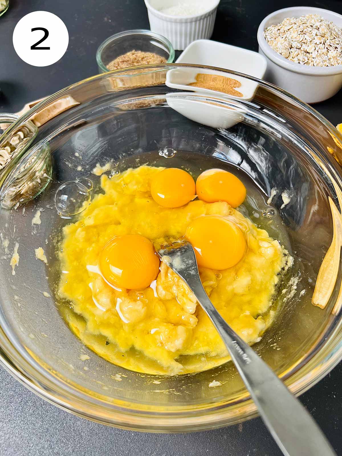 4 eggs added to the smashed bananas in a glass bowl with a metallic fork inside.