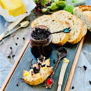 Elderberry jam, and a few slices of bread on a wooden board and a piece of sourdough bread with butter and jam.