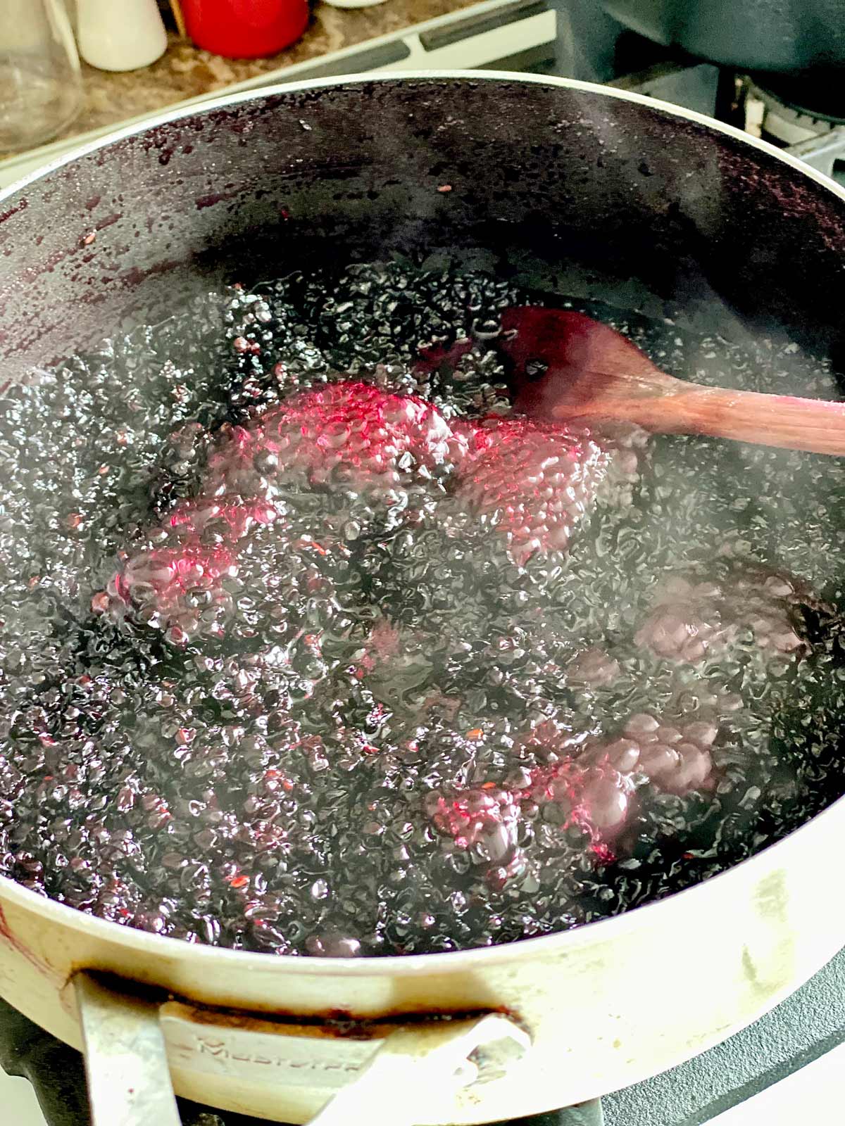 Elderberry jam bubbling away in a grey pan witha wooden spoon tucked in.