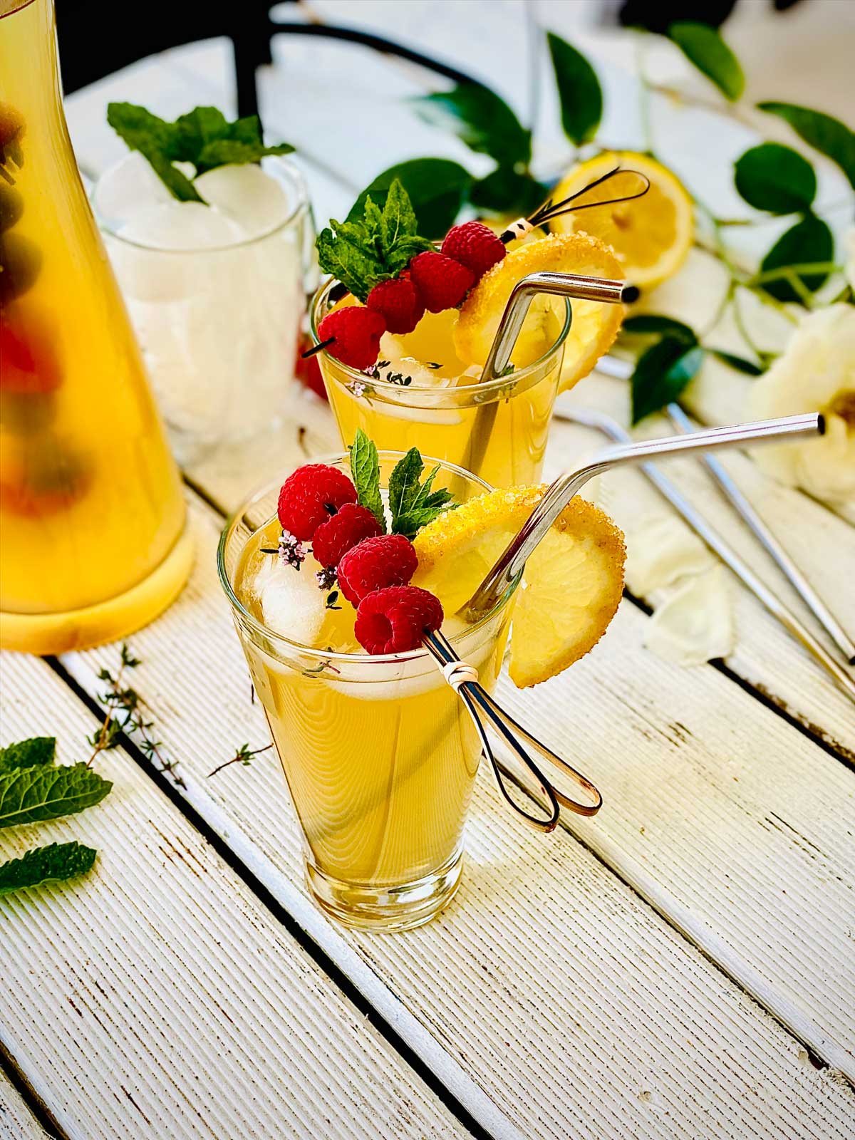 Two fancy glasses of elderflower cordial, decorated with sugar glacéd lemon slices and raspberry skewers and mint leaves with metallic straws tucked in
