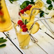 Two fancy glasses of elderflower cordial, decorated with sugar glacéd lemon slices and raspberry skewers and mint leaves with metallic straws tucked in