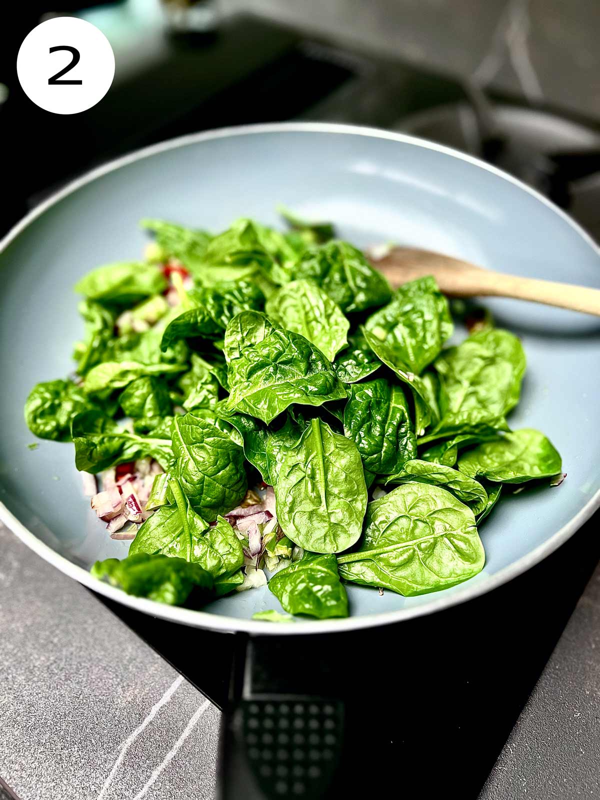 Fresh Spinach added to the sautéed vegetables in a frying pan with a wooden spatula in.