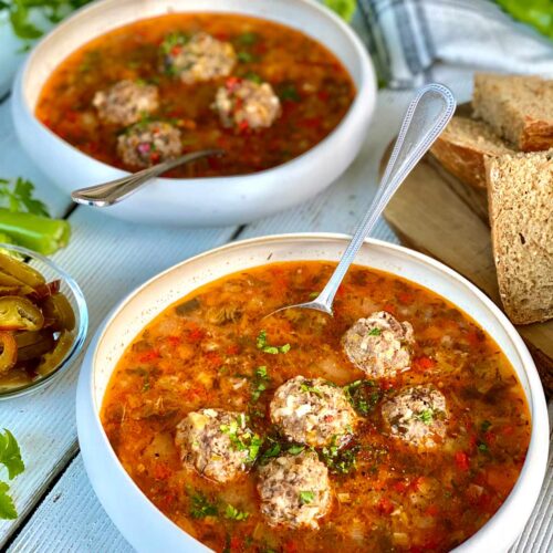 2 meatballs soup bowls, a glimpse of wholemeal bread slices on a chopping board on the right a kitchen towel and green peppers and fresh herbs in the far end