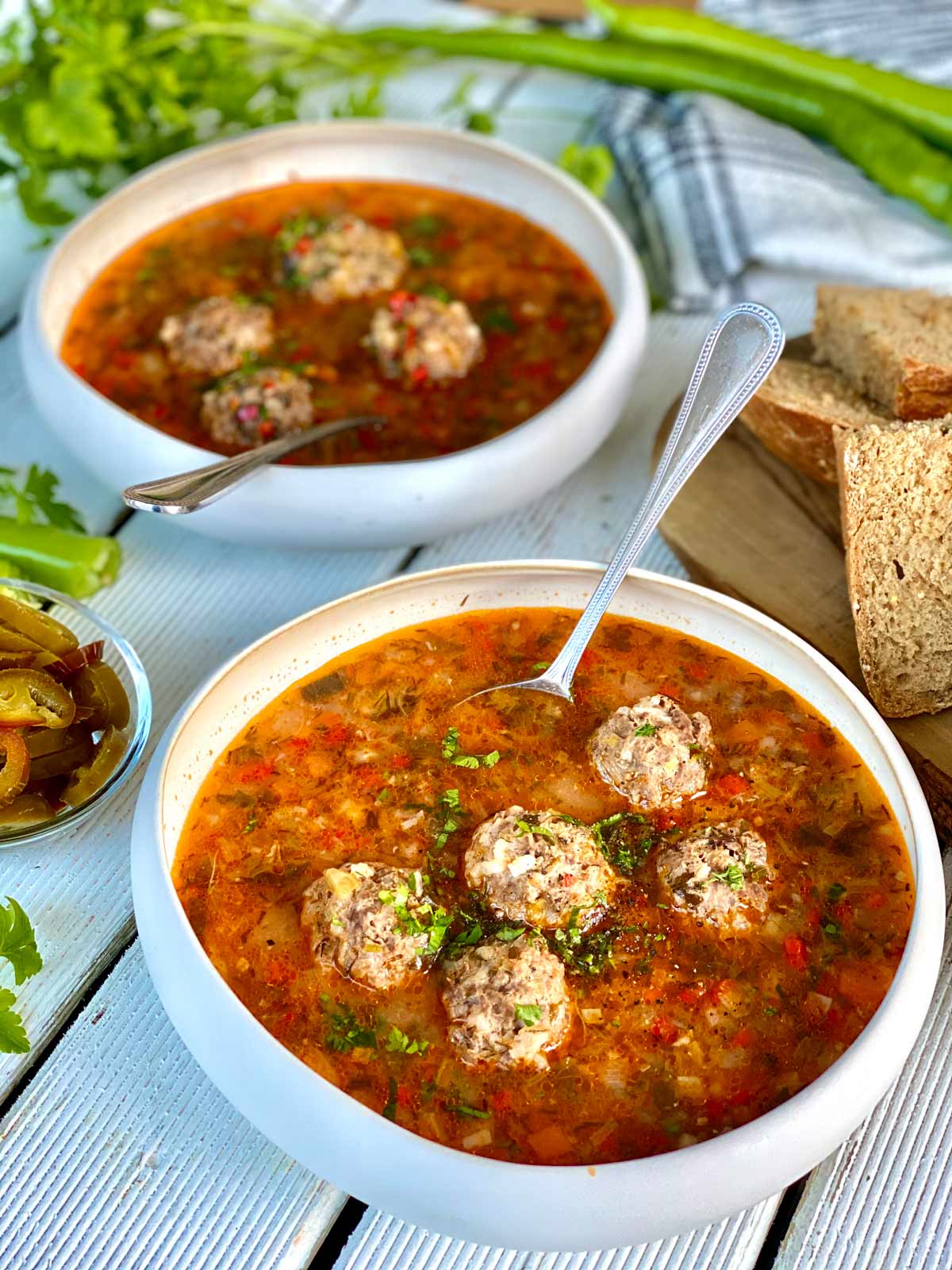 2 meatballs soup bowls, a glimpse of wholemeal bread slices on a chopping board on the right a kitchen towel and green peppers and fresh herbs in the far end