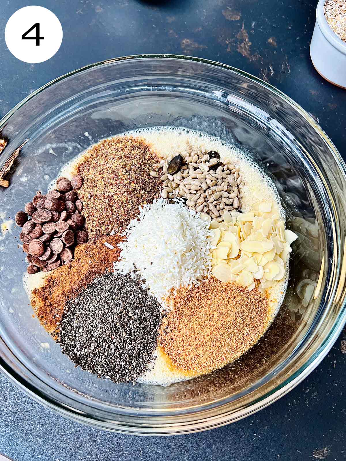 A transparent glass bowl containing beaten eggs, bananas, chocolate chips, flakes, sunflower, and pumpkin seeds, flaked almonds, desiccated Coconut, cinnamon, Chia seeds, and dried dates powder.