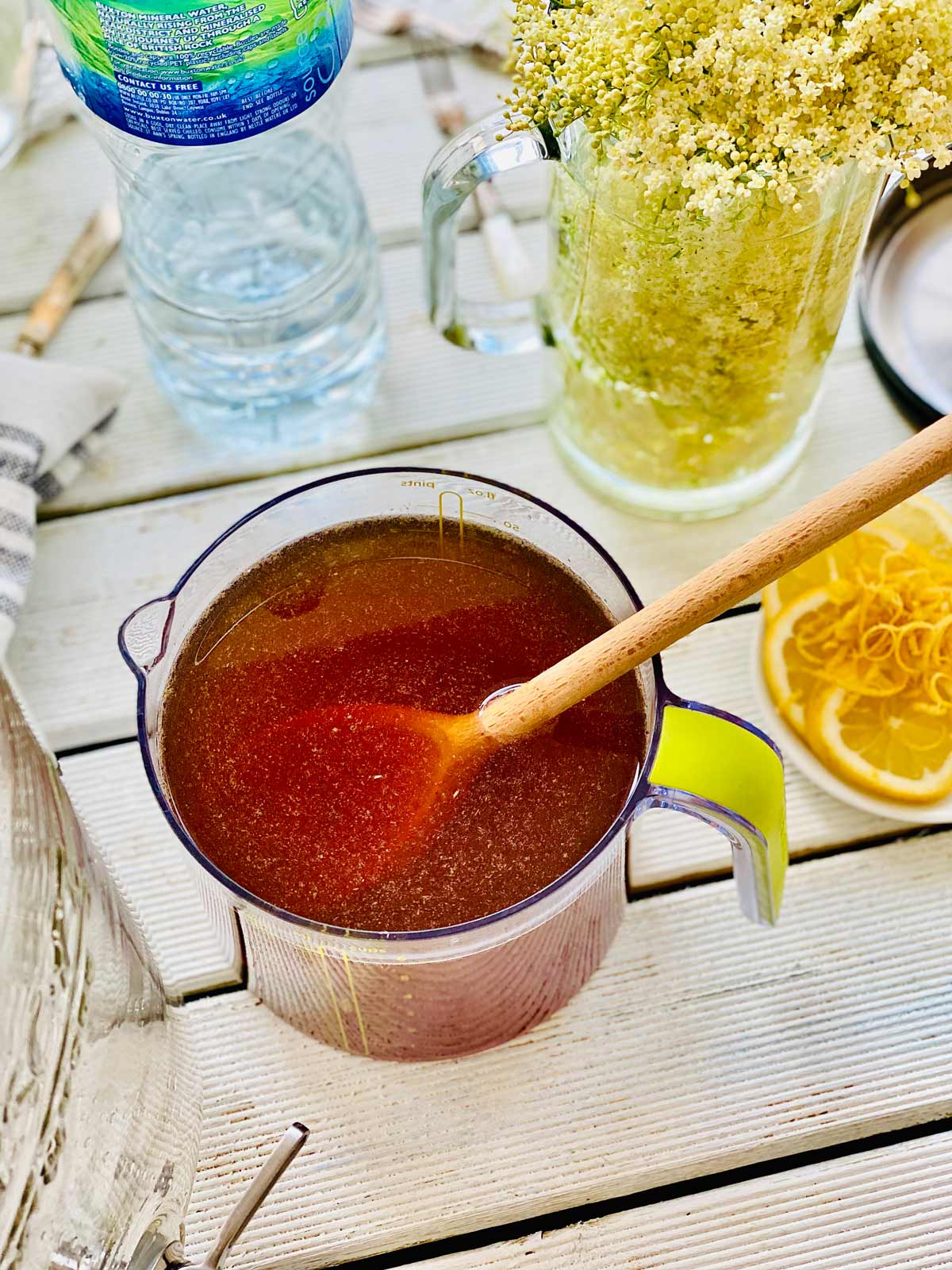 Water mixed with sugar in a jug with a wooden spoon and a jug with elderflower tucked in and lemon slices with lemon rind on the whiteboard