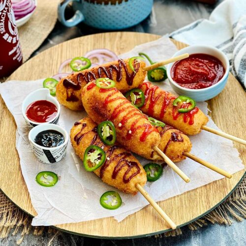 Korean corn dog on a wooden plate with a zigzag drizzle of sriracha and brown sauce and jalapeño slices scattered over