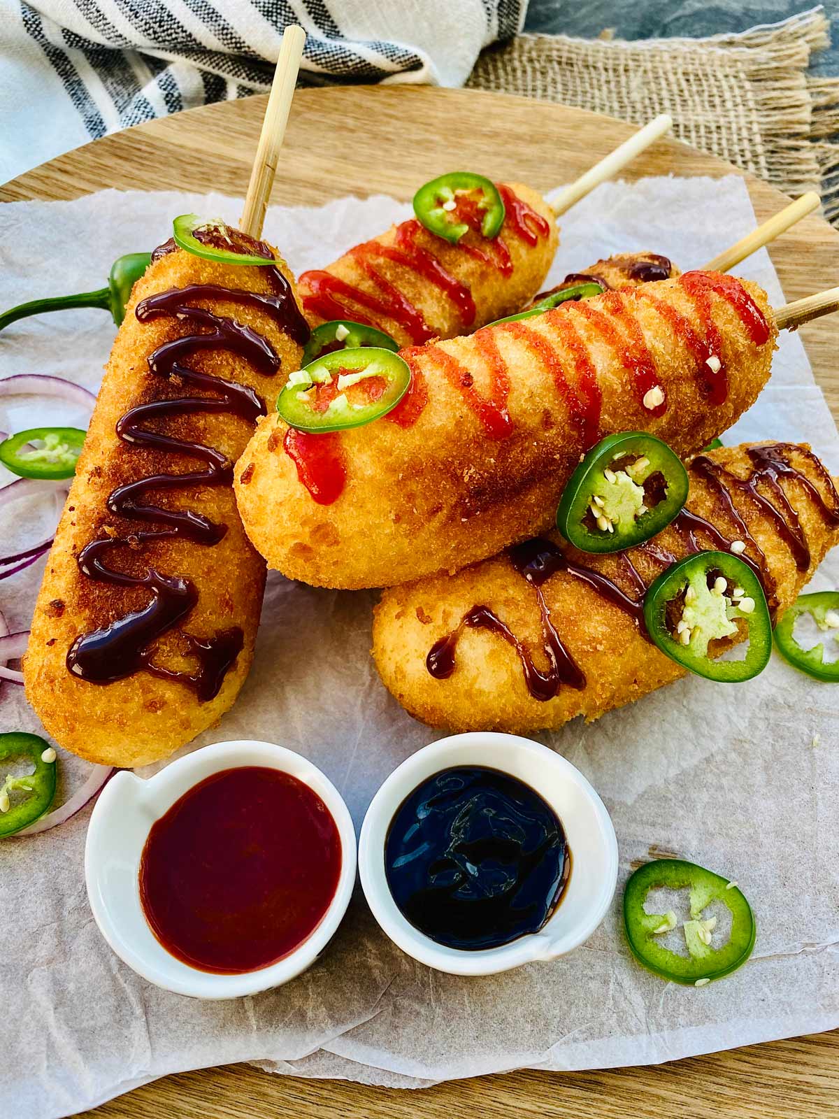 Korean corn dogs on a wooden plate with two tiny white serving dishes filled with red and brown sauces