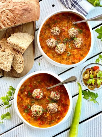 two white bowls with meatballs soup and spoons in with bread slices on the left and a small serving dish on the right filled with pickled jalapenos some fresh parsley leaves scattered around and a green pepperright
