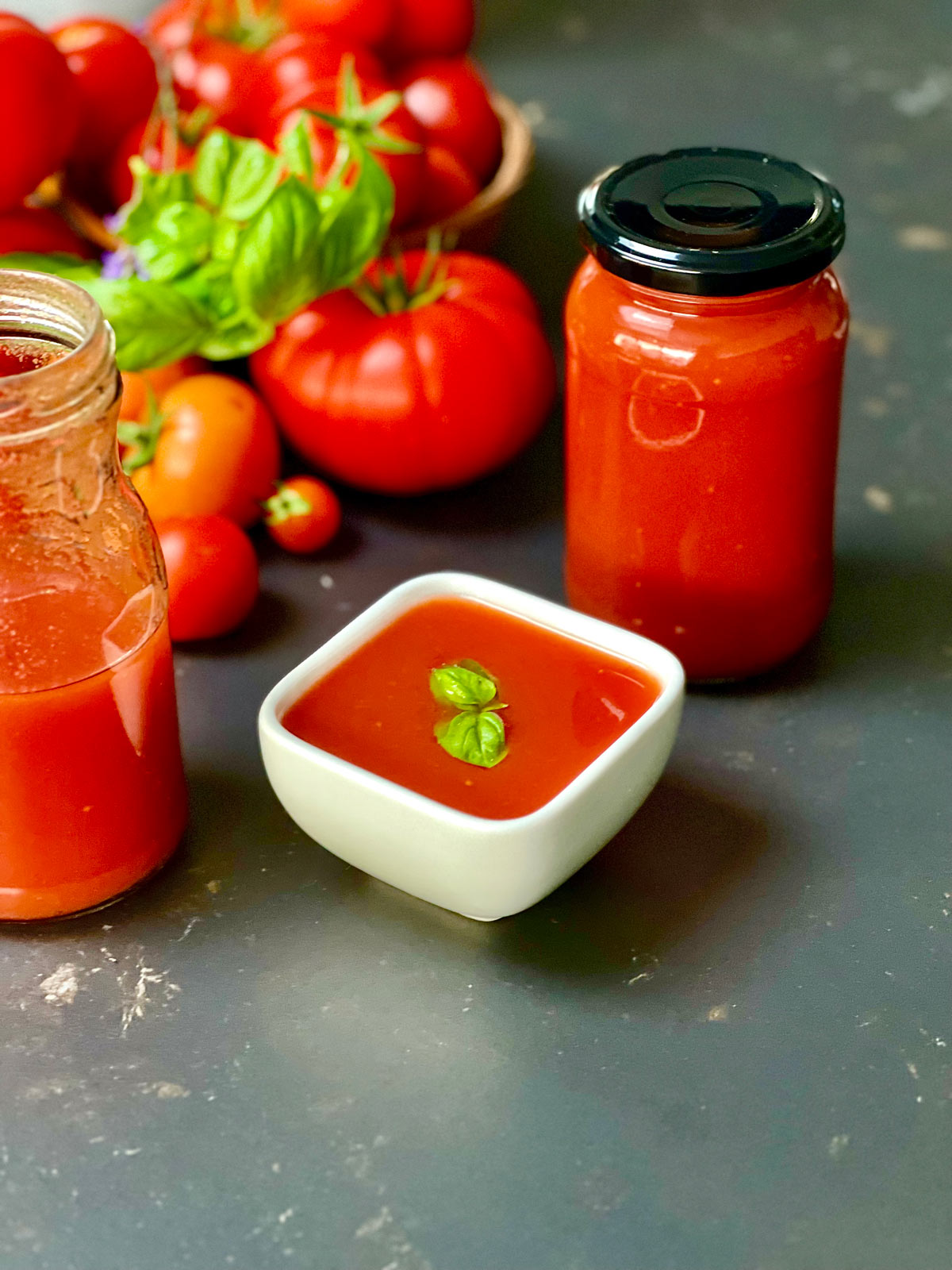 Half full jar to the left, fresh tomatoes and basil further up, a passata jar full to the right and a small dish containing passata si pomodoro and a basil leaf.