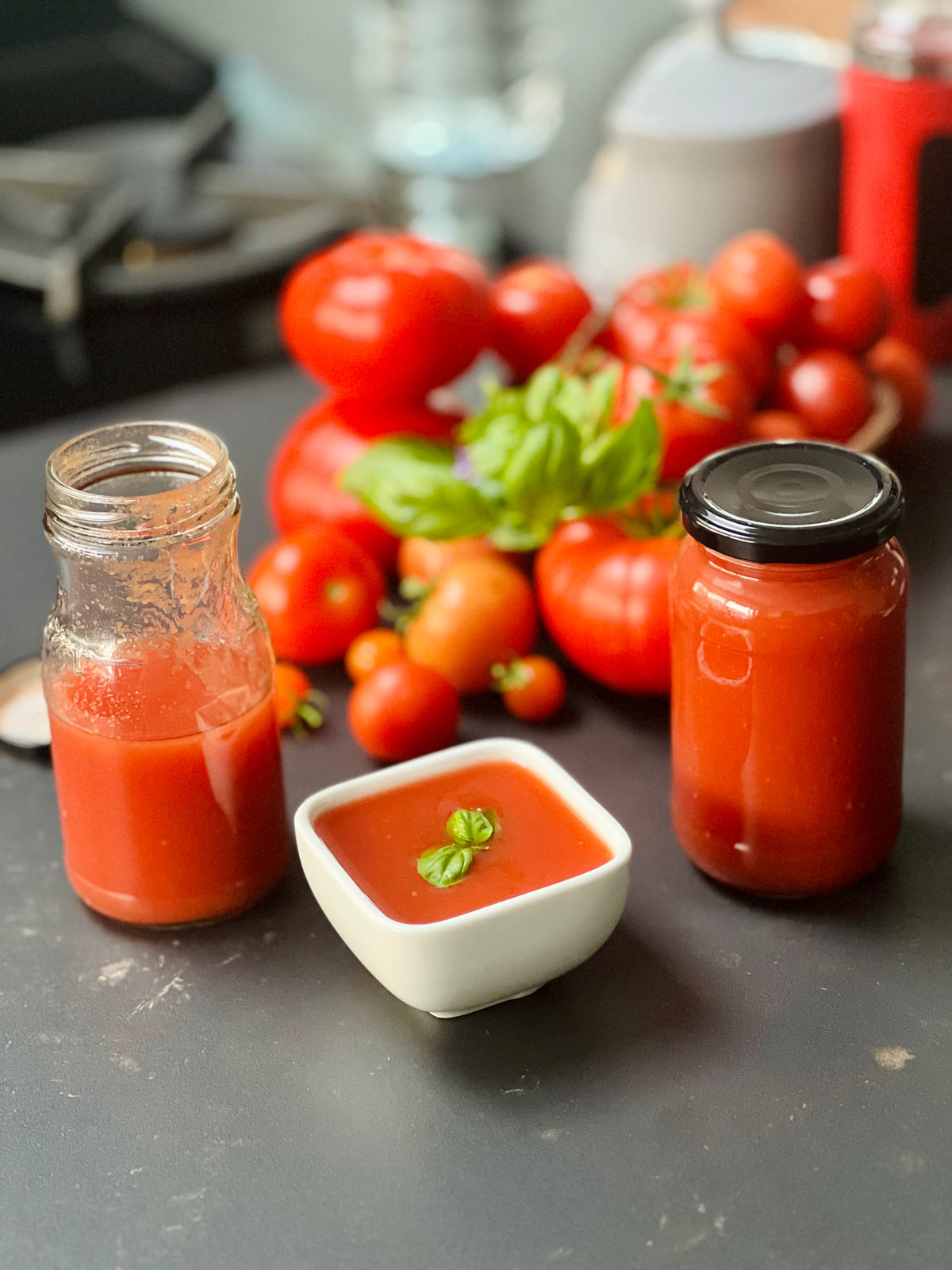 Two jars, set aside, one full one halfway filled with passata, and the white ceramic dish in the middle containing tomato, passata and some basil leaves.
