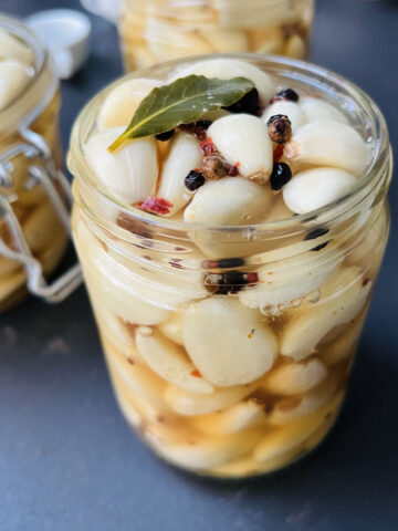 Pickled garlic jars on a dark gray worktop topped with spices and bay leaves.