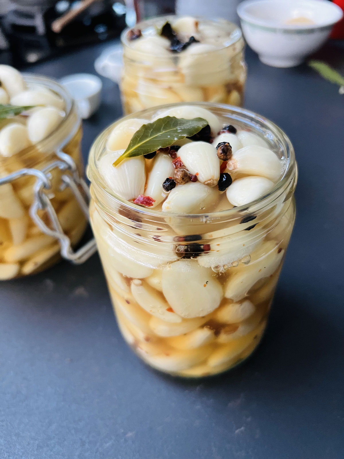 3 Pickled garlic jars on a dark gray worktop topped with spices and bay leaves.