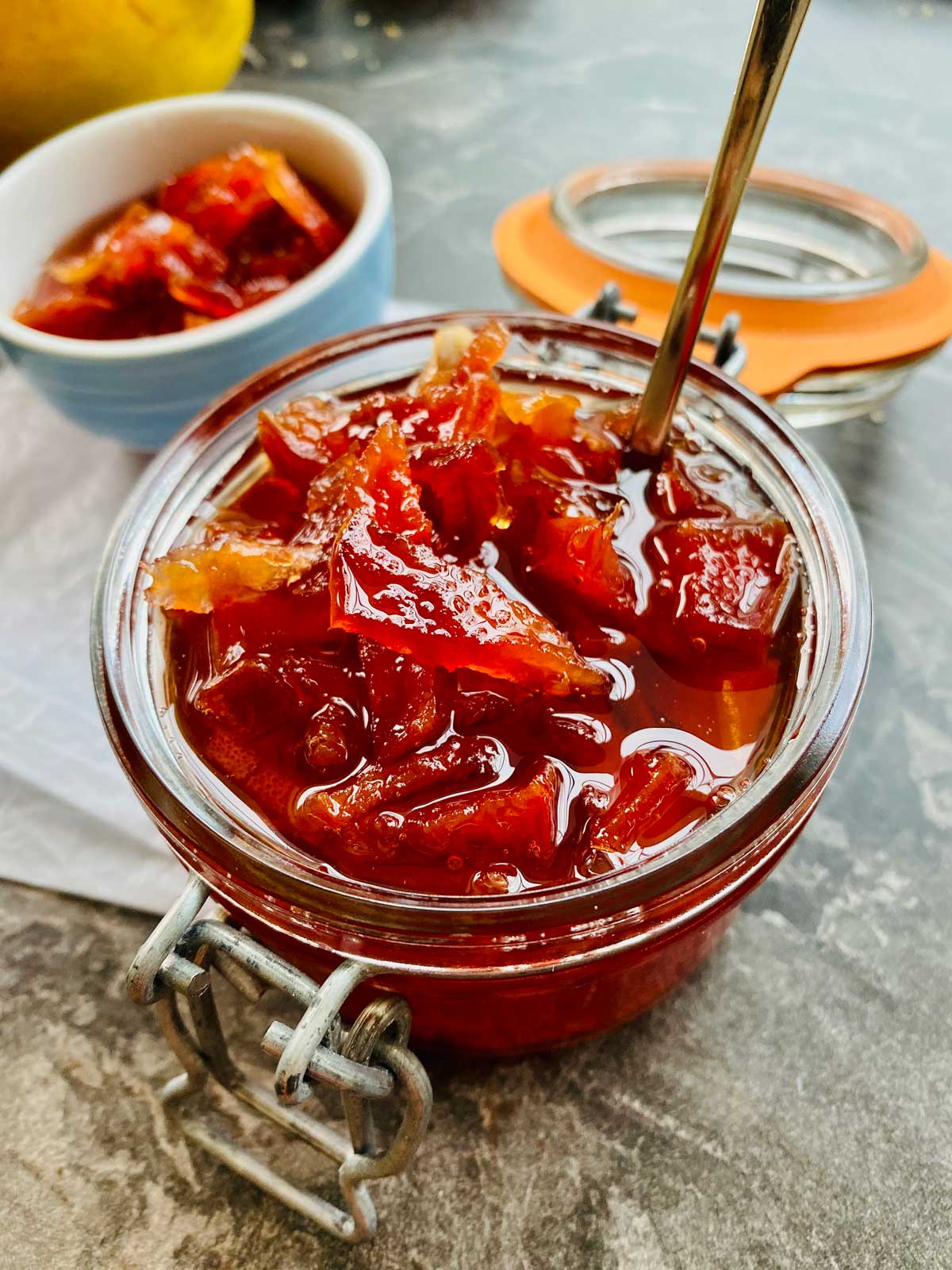 Close up of deep orange-red junky quince jam in a preserve jar with the lid opened and a teaspoon in