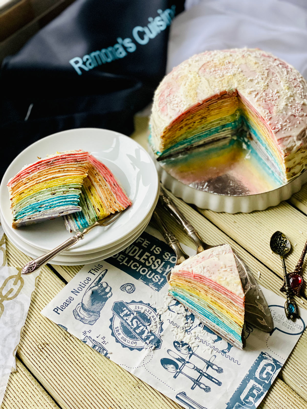 Two rainbow crepe cake slices on a stack of white plates and one slice closer on a patterned black and white paper