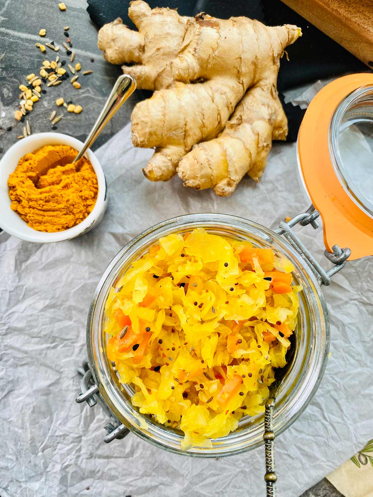 Raw sauerkraut with turmeric in a jar on grey creased paper and turmeric in a small white ceramic dish to the right and a fresh ginger root right at the top